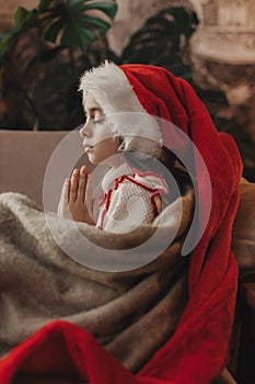 Close-up Caucasian child, lovely baby girl in Santa hat, makes cherished wish and cute presents, surrounded by Christmas