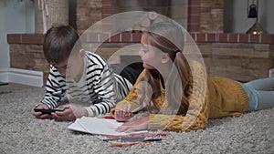 Close-up of Caucasian cute girl coloring with crayons and looking at her brother playing games in smart phone. Siblings