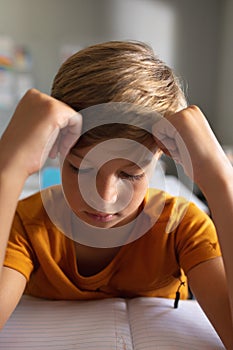 Close-up of caucasian elementary schoolboy with head in hands studying at desk in classroom