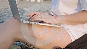 Close up of Caucasian gir working with laptop, typing on computer keyboard on the beach by the sea during sunset. Woman