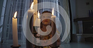 Close-up of Caucasian girl`s face behind candles. Weird child holding doll and looking around. Scare, fear, strangeness.