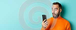 Close-up of caucasian man staring at phone screen with surprised face, wearing orange sweater, light blue background