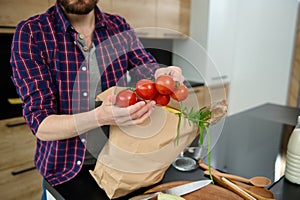 Close-up of a Caucasian man taking out vegetables and healthy food from a paper eco bag, holding a sprig of fresh tomatoes in his