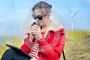Close up Caucasian woman hold a cup of coffee and drink near meadow and wind turbines on the background