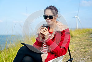 Close up Caucasian woman hold a cup of coffee and look at the camera and sit near meadow and wind turbines on the background