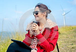 Close up Caucasian woman hold a cup of coffee and look to left side and sit near meadow and wind turbines on the background