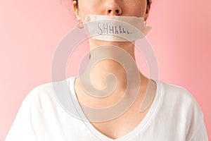 Close up of caucasian woman& x27;s face with her mouth taped shut. Tape has the inscription Shhh. Pink background photo