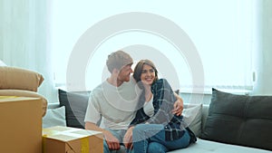 Close up of Caucasian young happy man and woman sitting on sofa in new house with many cardboard boxes on moving day