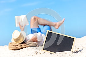 Close up of a chalkboard on the sand of a beach, background happy smiling caucasian tourist asian young man relax and reading book