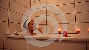 Close-up of a charming African American woman taking a relaxing bubble bath by the light of scented red candles. Cleanliness, hygi