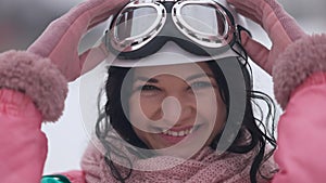 Close-up of charming joyful woman looking at camera with toothy smile putting on ski goggles in slow motion. Portrait of