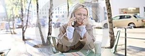 Close up of cheerful blond girl, woman talking on mobile phone, sitting in city centre, outdoor cafe, taking break from