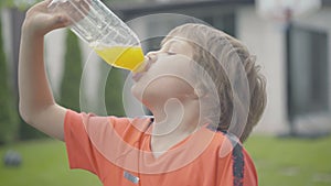 Close-up of cheerful Caucasian boy drinking healthful juice outdoors. Portrait of happy kid enjoying refreshing drink at