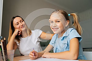 Close-up of cheerful primary daughter doing homework with young mother sitting at home table by window, looking at