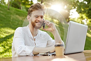 Close up of cheerful red-haired guy with trendy hair and beard sitting outside, working on laptop computer, talking on