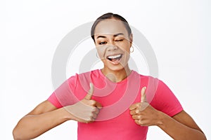 Close up of cheerful woman winks and shows thumbs up, being supportive, recommends smth, praise and compliments, stands
