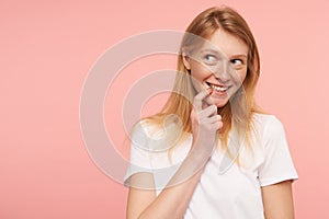Close-up of cheerful young attractive female with loose raising hand to her face while looking cunningly aside with light smile,
