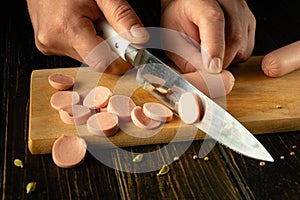 Close-up of a chef hands with a knife slicing vienna sausage on a kitchen board. Low key concept of preparing a delicious