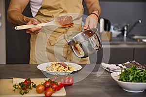 Close-up chef pouring tomato sauce on Italian pasta, plating up the dish before serving. Man cooking dinner at home