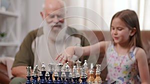 Close-up chess board with blurred curious girl talking to senior man sitting at background. Positive grandfather