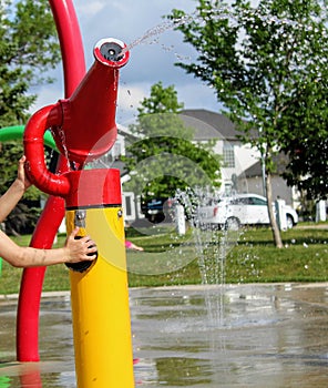 Close-up of a child's hand presses the sprinkler button at the children splash pad. A small fountain in the background.