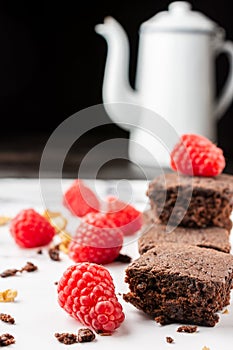 Close-up of chocolate brownie with crumbs, walnuts, red raspberries and white coffee pot, selective focus, on white marble table,