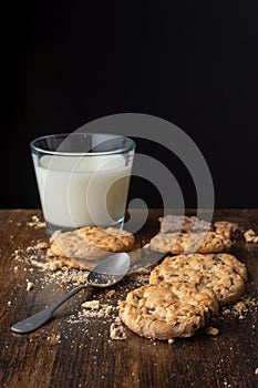 Close-up of chocolate chip cookies, crumbs, spoon and glass of milk, with selective focus, on wooden table and black background