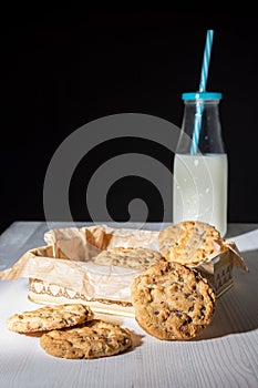 Close-up of chocolate chip cookies, metal box and bottle of milk with blue straw, on white wooden table and black background