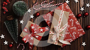 Close-up of Christmas gifts lie on a brown wooden table among holiday decor, fir trees, balls, stars and striped candies, top view