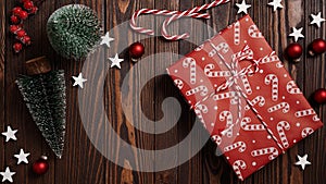 Close-up of Christmas gifts lie on a brown wooden table among holiday decor, fir trees, balls, stars and striped candies, top view