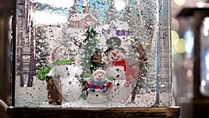 Close-up of Christmas glass snow globe with a family of snowmen and falling snow from glitter. Slow motion.