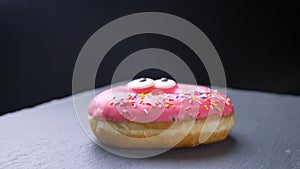 Close-up circling around shot of delicious glazed pink donut with multicolored chips and eyes spinning slowly on gray