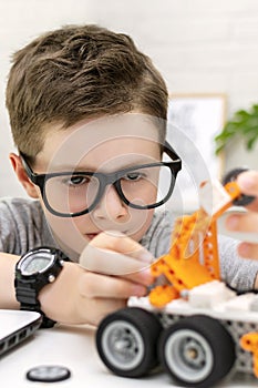 Close-up of a clever boy in eyeglasses builds and programs a robot car at home. The child is learning coding and