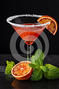 Close-up of cocktail glass with blood orange martini and sugar, with half orange and mint, on black background