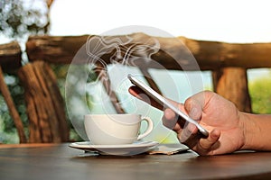 Close up coffee cup with hands of young man holding mobile phone in nature background. Selective focus and shallow depth of field.