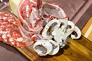 Close up composition of cheese, bacon, salami and white sliced mushrooms on a wooden board