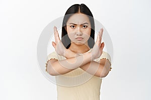 Close up of confident and serious asian girl shows cross sign, stop and prohibit gesture, standing in tshirt over white