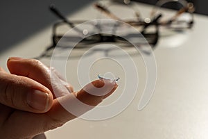 Close-up of a contact lens on a female index finger against the background of glasses on a white table.