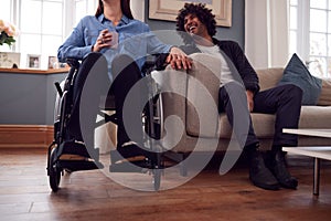 Close Up Of Couple At Home With Disabled Woman In Wheelchair Holding Hands With Man Sitting On Sofa