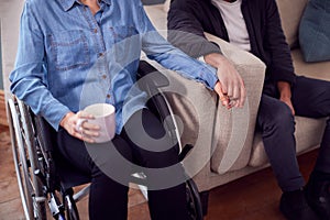 Close Up Of Couple At Home With Disabled Woman In Wheelchair Holding Hands With Man Sitting On Sofa