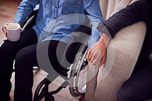 Close Up Of Couple At Home With Disabled Woman In Wheelchair Holding Hands With Man Sitting On Sofa