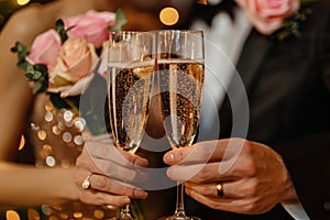 Close-up of a couple& x27;s hands holding champagne flutes in a toast, surrounded by soft pink roses