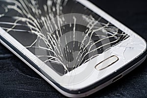 Close-up of a cracked touchscreen of a white phone. Broken screen glass of a smartphone on the black wooden background.