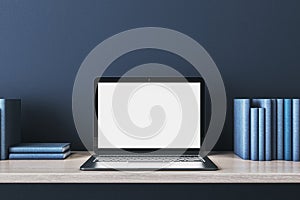 Close up of creative wooden designer desk with books and empty white mock up laptop screen on blue concrete wall.