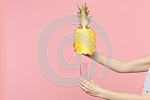 Close up cropped photo of female hold in hands half of fresh ripe pineapple fruit, glass cup  on pink pastel