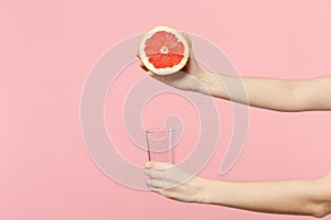 Close up cropped photo of female holding in hands half of fresh ripe grapefruit, glass cup isolated on pink pastel wall