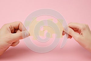 Close up cropped photo of hands holding trying to connect couple wooden jigsaw puzzle pieces on pastel pink