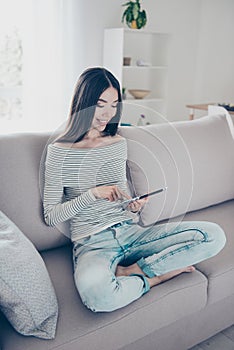 Close up cropped photo of young cheerful chinese lady browsing on her tablet, sitting on beige couch indoors at home, wearing casu