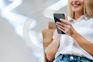 Close-up cropped shot of unrecognizable cheerful blonde young woman using mobile phone, holding shopping paper bags with