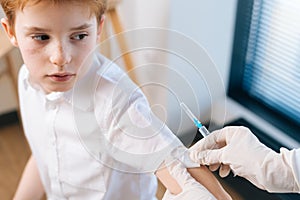 Close-up cropped shot of unrecognizable female doctor vaccinating little child boy on shoulder at hospital by window.